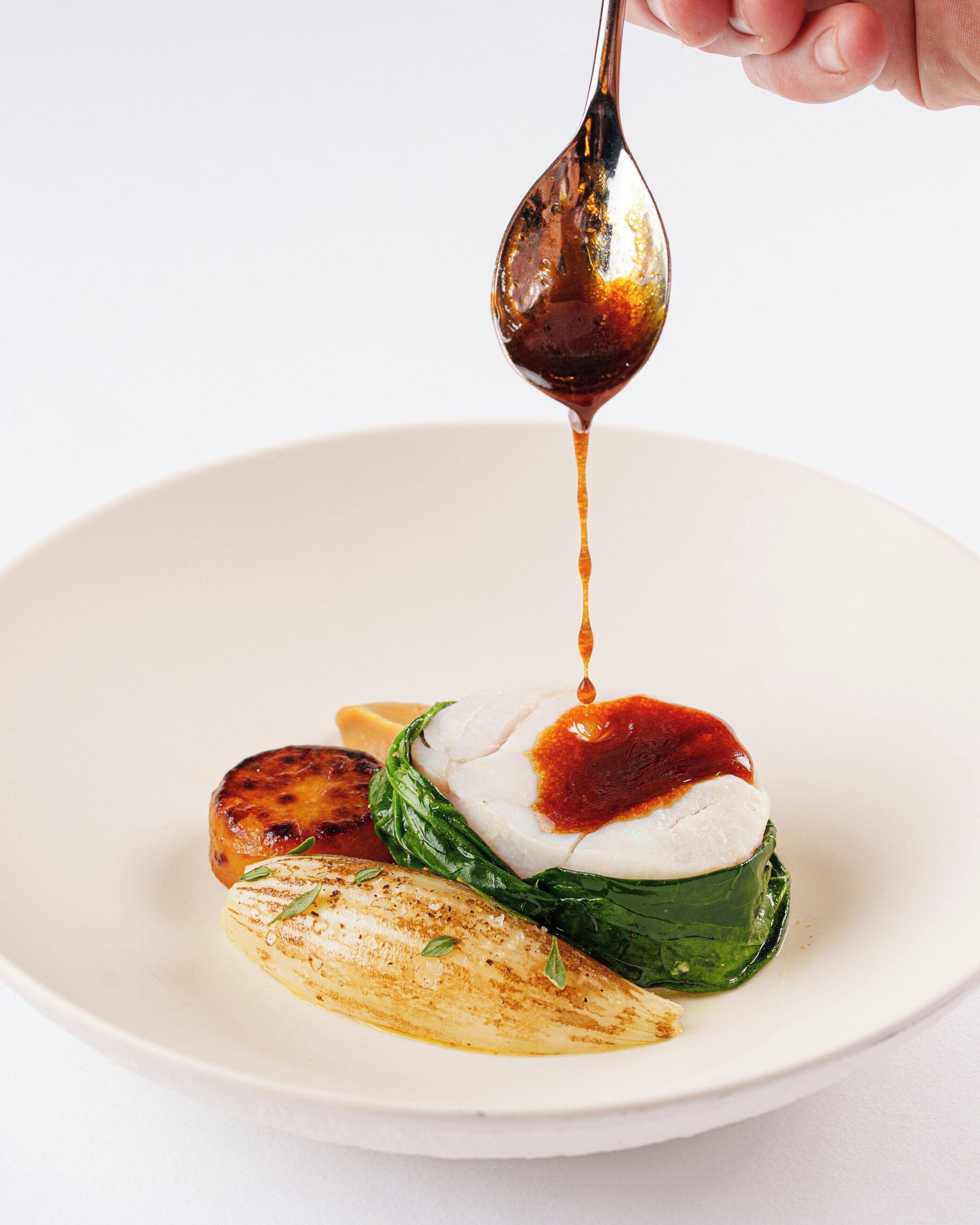 A white plate with a grilled meat, a poached egg, and a green vegetable, with sauce being poured over the egg.