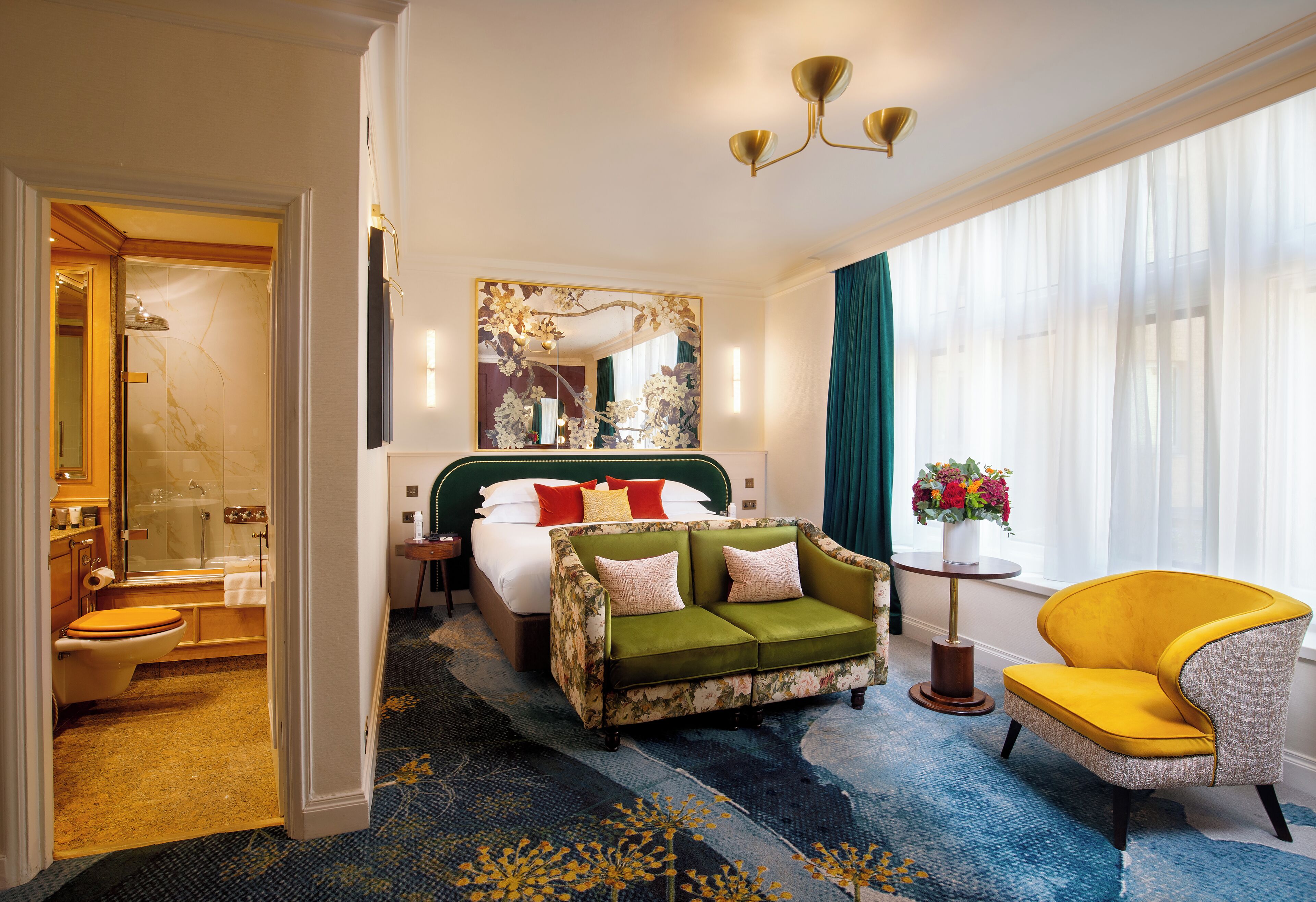A hotel room with a bed, a green sofa, a yellow armchair, a bathroom, and a large window with curtains.