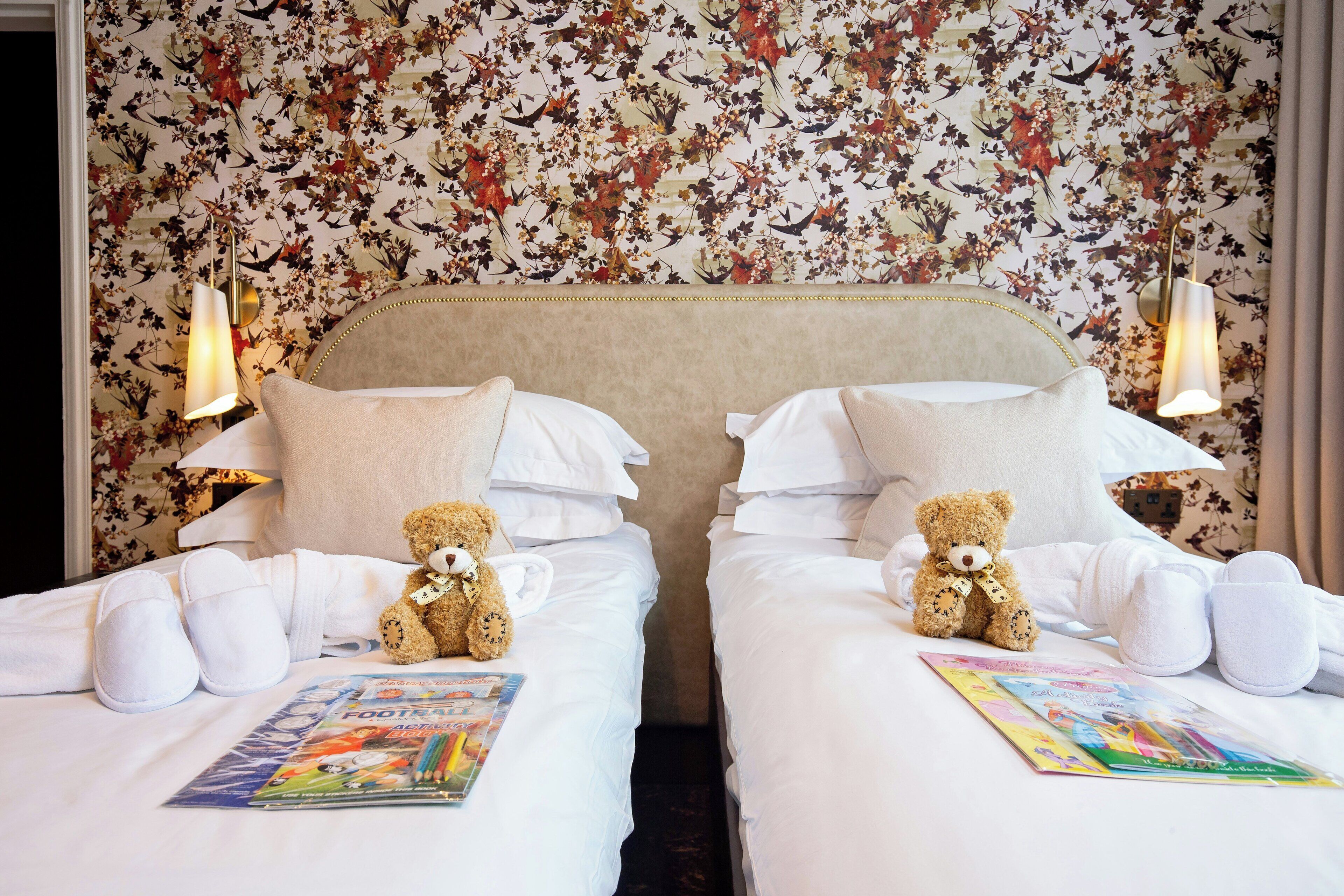 A hotel room with two beds, floral wallpaper, white bedding, and two teddy bears.