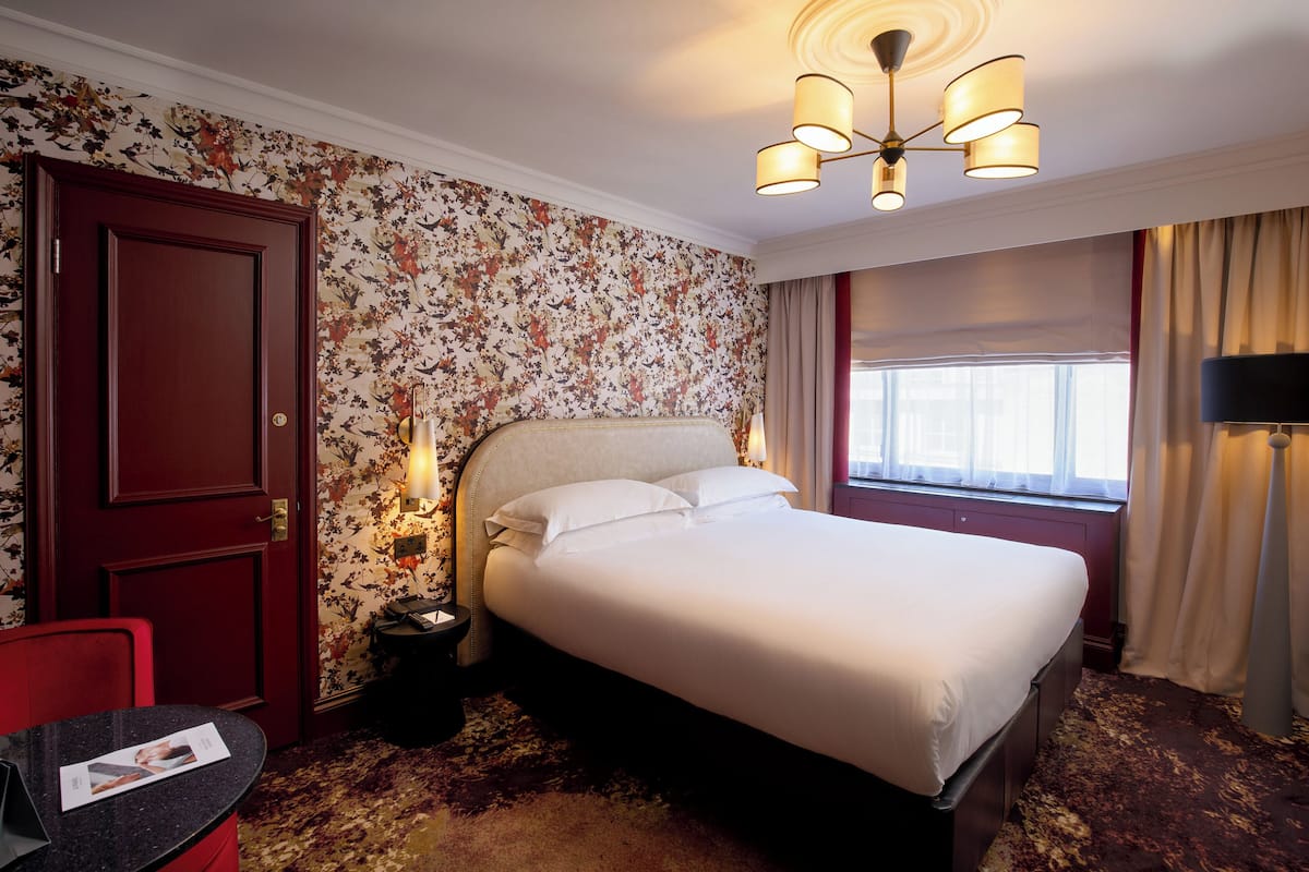 Een hotelkamer met een groot bed, een bureau met stoel, een nachtkastje, bloemenbehang, een kroonluchter en een raam met gordijnen.