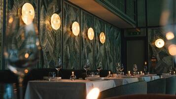 Interior de restaurante con mesas dispuestas para comer, destacando un muro con diseño y luminarias colgantes.