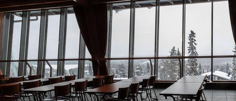Ein Essbereich mit Tischen und Stühlen, große Fenster bieten Blick auf schneebedeckte Bäume und ein Gebäude in der Ferne.