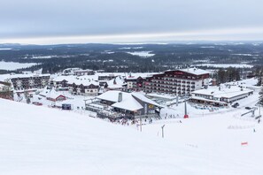 A ski resort with multiple buildings, including a large hotel with a red roof, surrounded by snow-covered terrain and distant mountains.