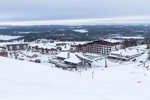 A ski resort with multiple buildings, including a large hotel with a red roof, surrounded by snow-covered terrain and distant mountains.