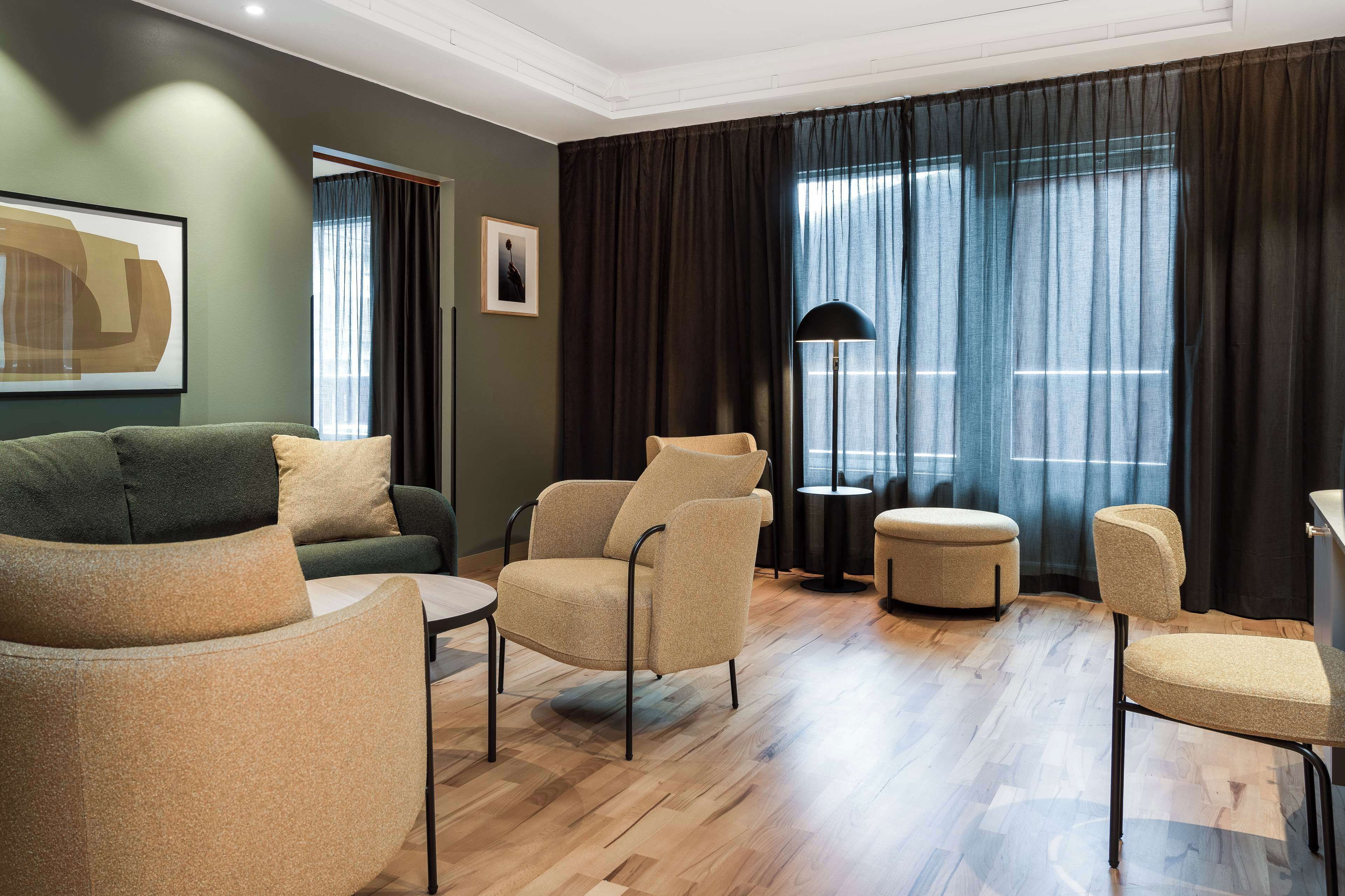 Una sala de estar moderna con un sofá verde, sillones color beige y un otoman redondo. Hay una lámpara de pie y un cuadro en la pared.