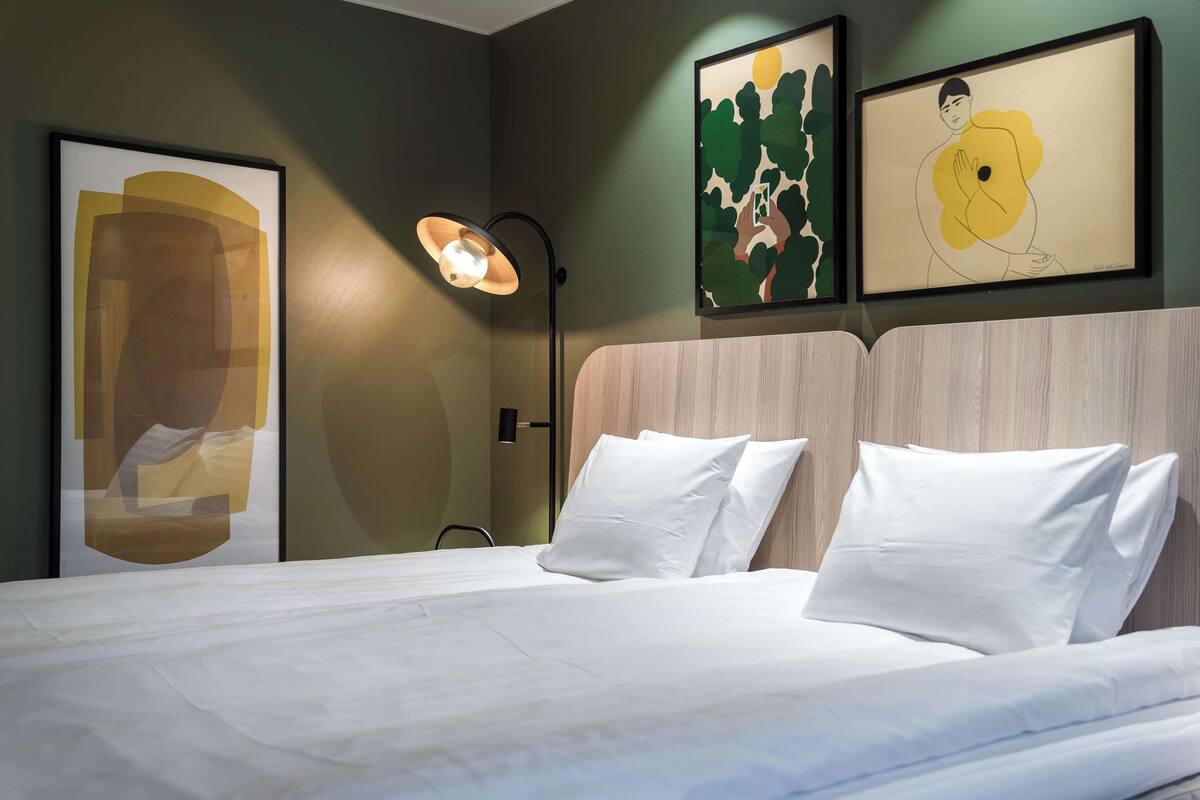 Ein ordentlich bezogenes Bett mit weißen Leinen, eine moderne Stehlampe und abstrakte Kunstwerke an der Wand.