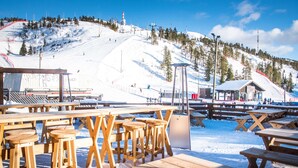 A wooden outdoor seating area with tables and stools, a small building, and a snowy mountain backdrop.