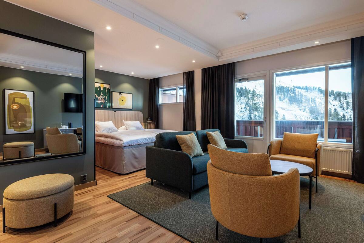Ein modernes Hotelzimmer mit Bett, Sofa, Sessel und Blick auf eine verschneite Berglandschaft.