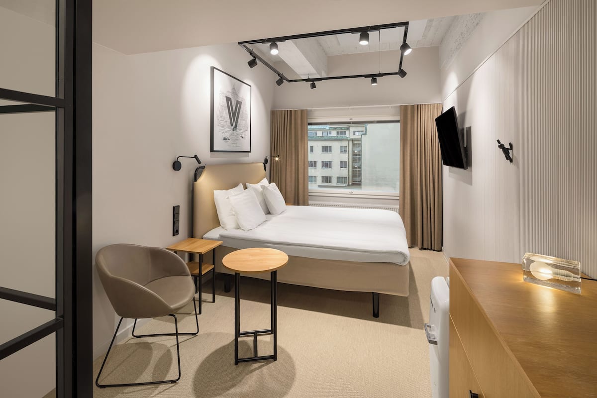 A modern hotel room with a bed, a chair, a small table, a TV, and a window with a view of buildings.