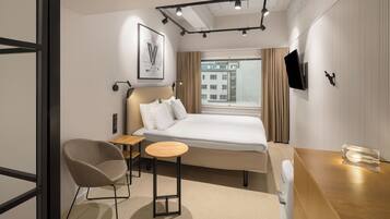 Habitación de hotel moderna con cama, una silla, una mesita, televisor y ventana con vista a edificios.