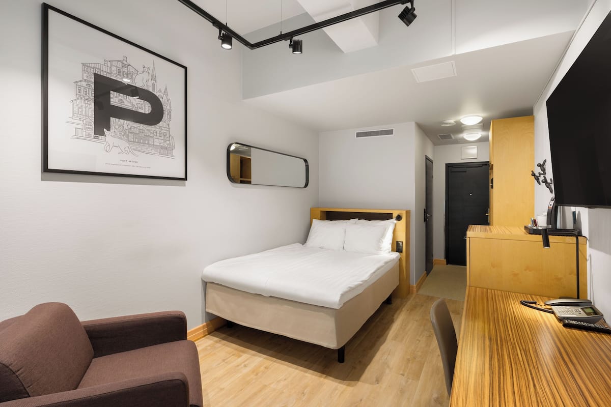 A hotel room with a bed, a chair, a desk with a phone, a mirror, and a framed picture on the wall.