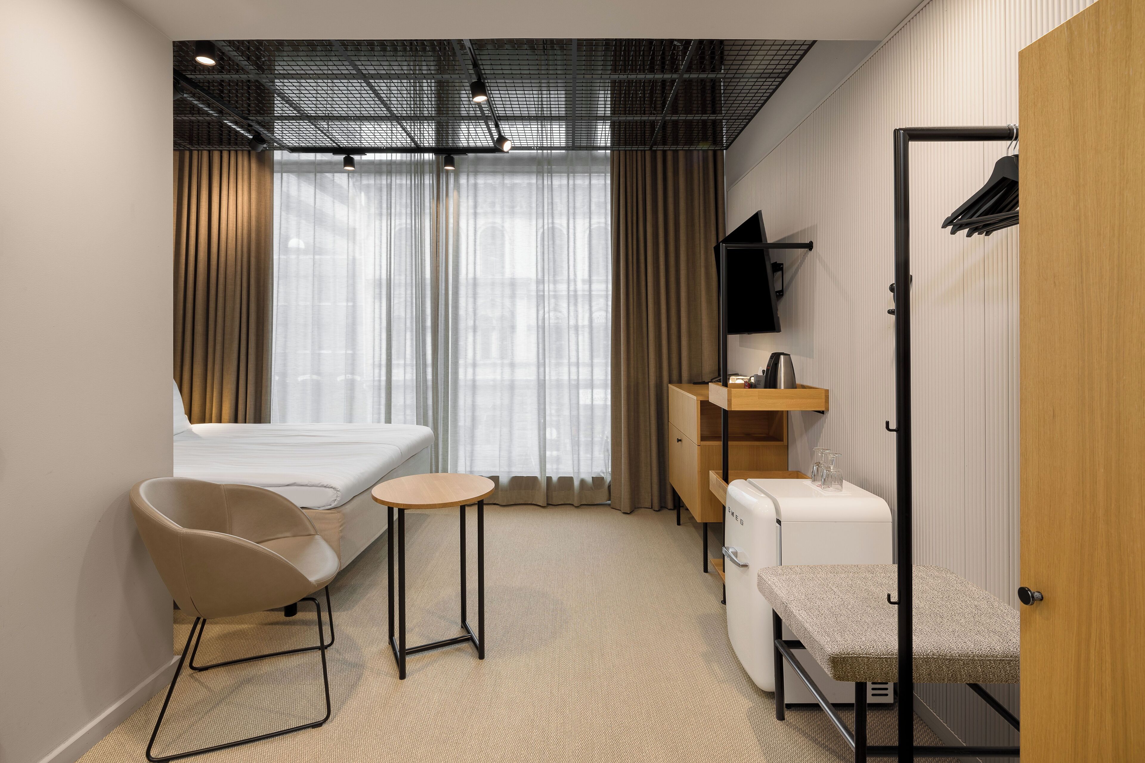 Habitación de hotel moderna con cama, una silla, una mesita, un escritorio, televisor y perchero.