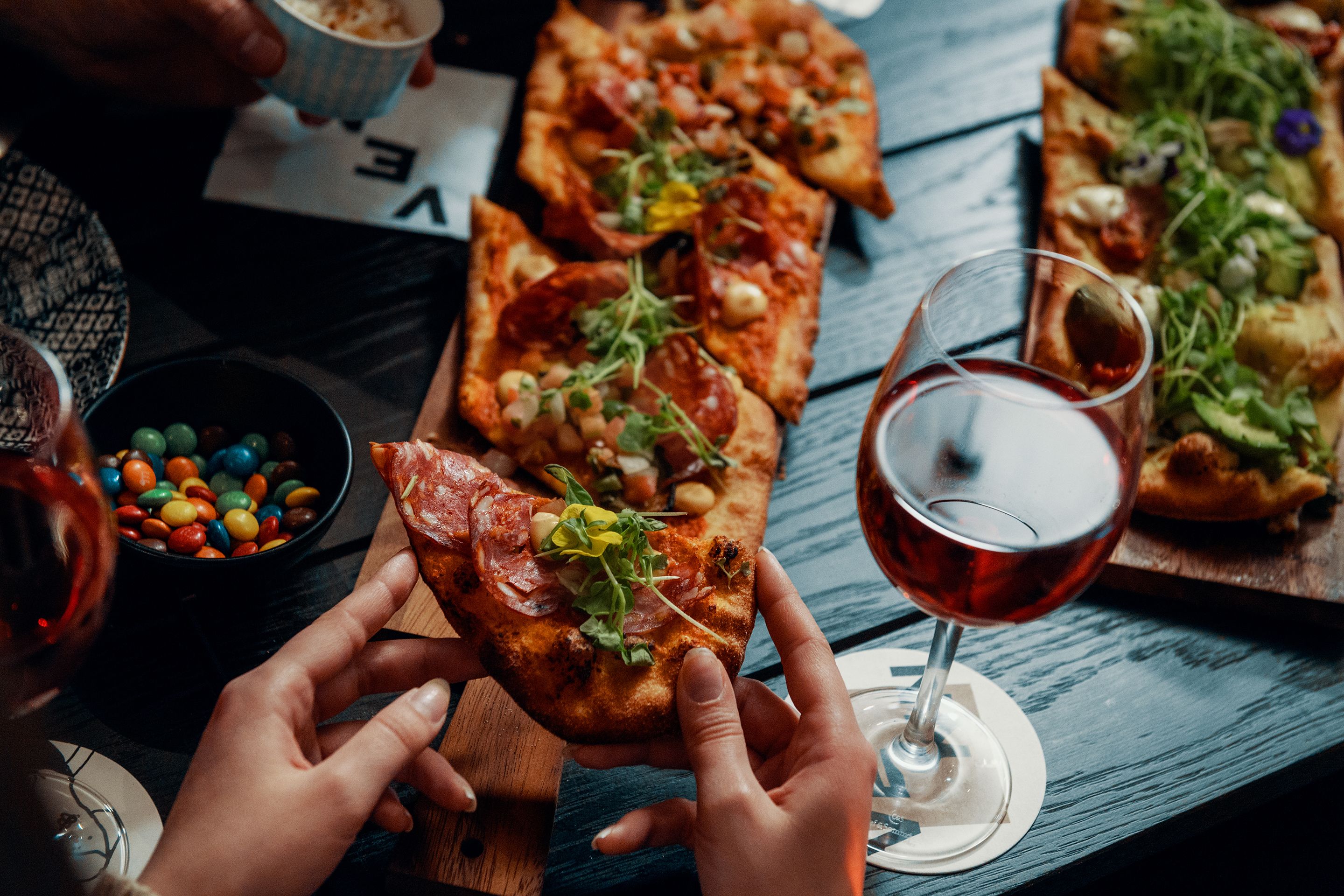 Ruokapöydällä on pizzaa, lasillinen viiniä ja kulho värikkäitä makeisia.