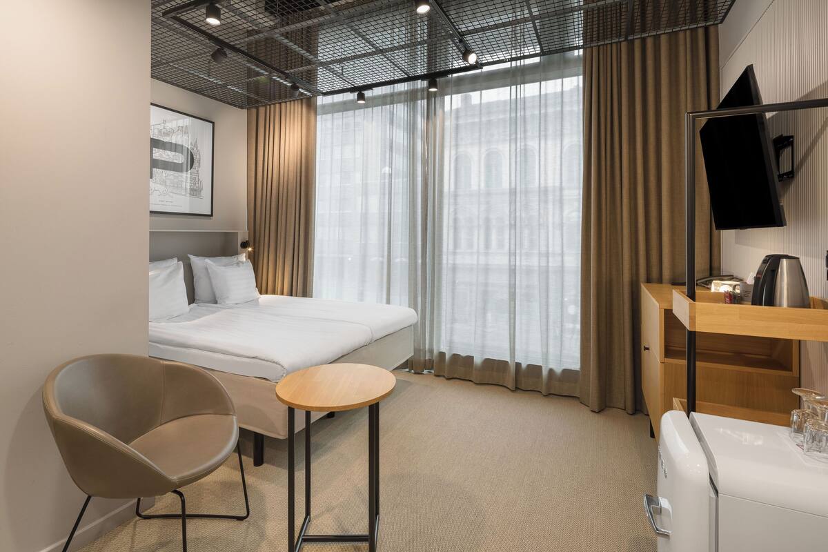 A modern hotel room with a bed, a chair, a small table, a desk, a TV, and a coffee machine.