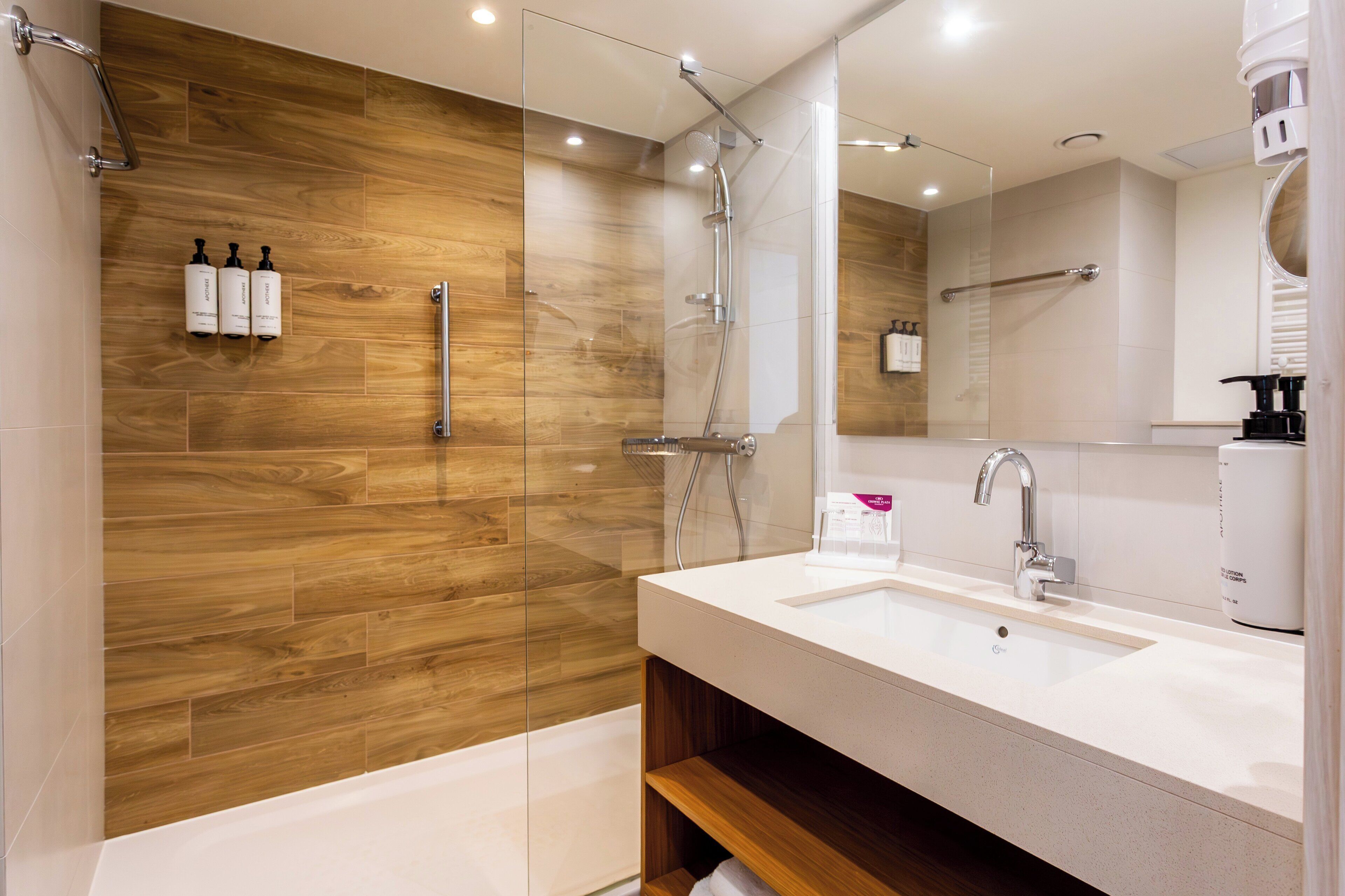 A modern bathroom with a wooden wall, a glass shower enclosure, a white sink, and a large mirror.