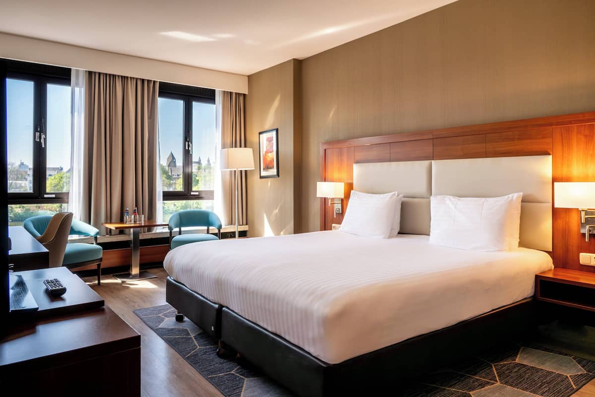Een hotelkamer met een groot bed, twee nachtkastjes met lampen, een bureau met stoel en uitzicht op het omliggende landschap door grote ramen.