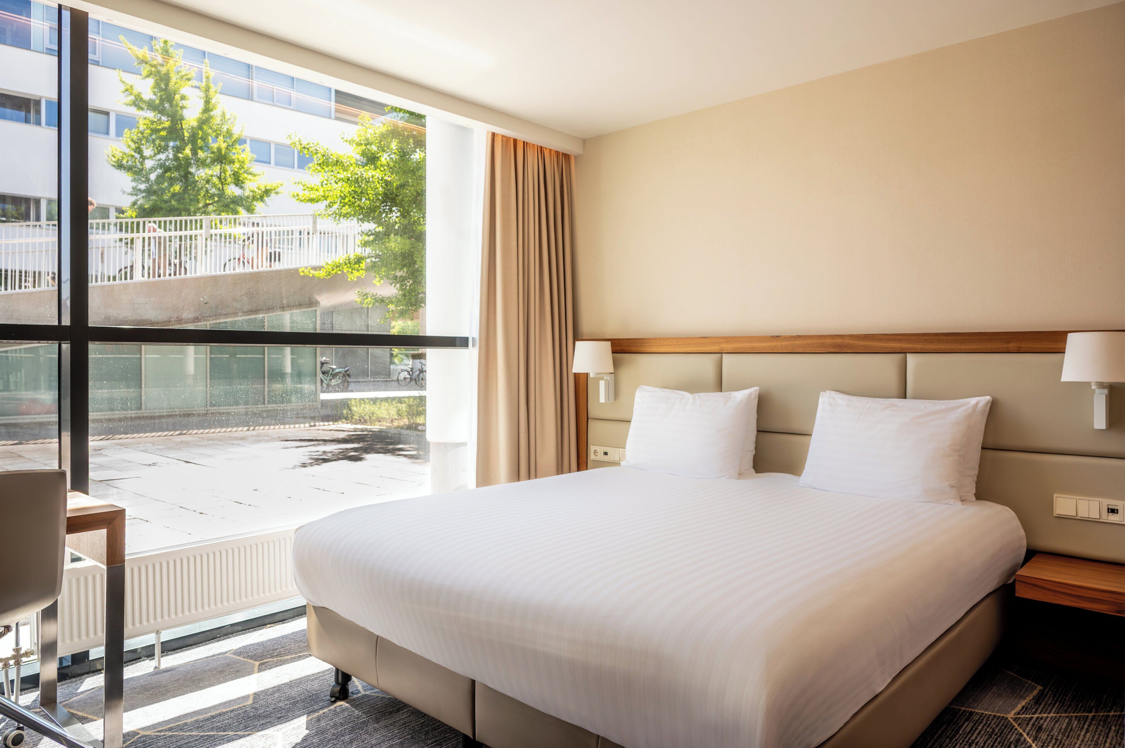 A hotel room with a large bed, a window with a view of a building and trees, a desk, and a chair.