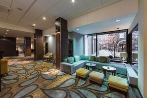 Un hall d’hôtel doté d’un tapis à motifs, d’un espace salon avec des canapés et des poufs, d’une grande fenêtre offrant une vue sur la ville et d’un comptoir de réception.
