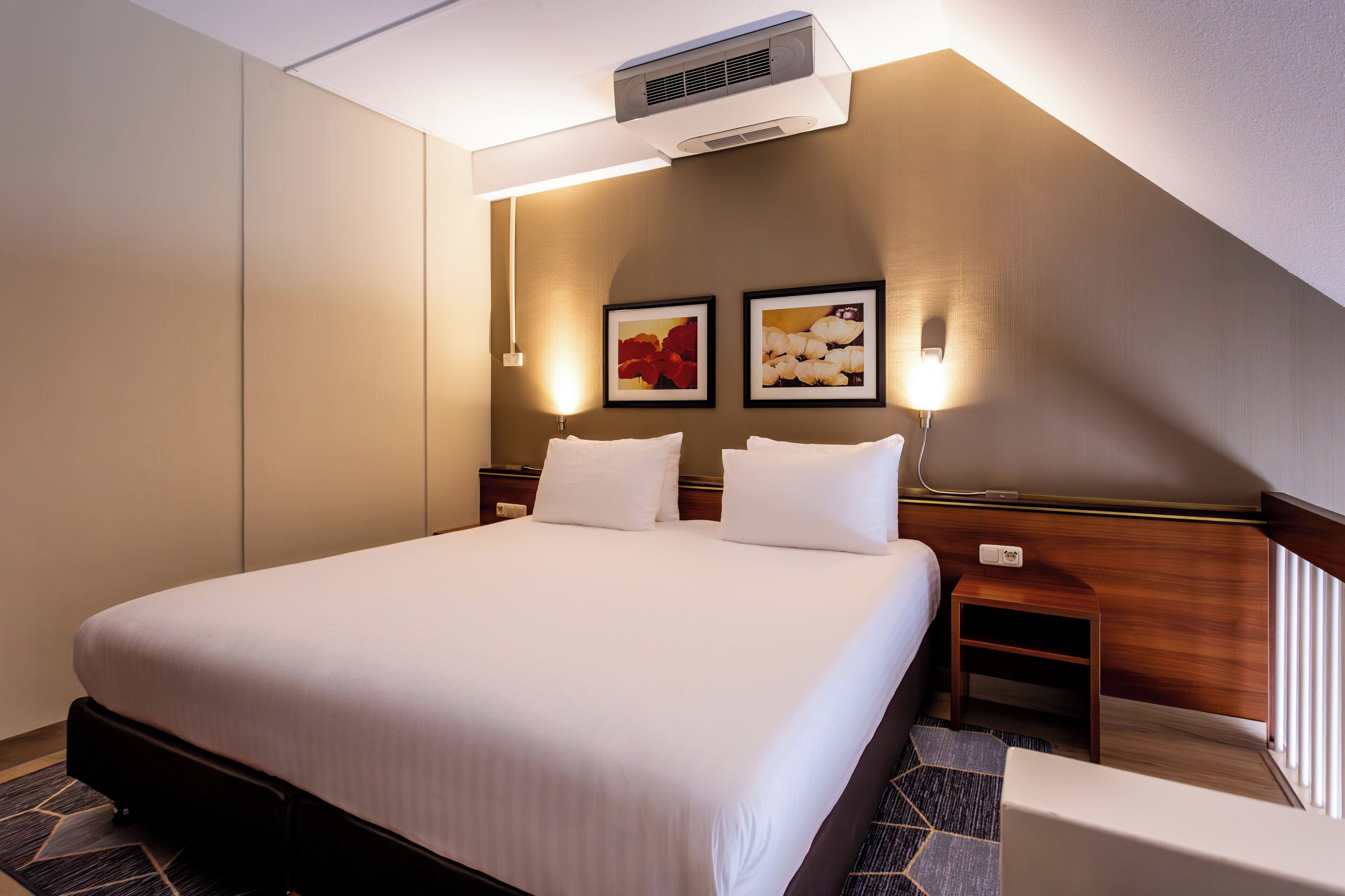 Ein Hotelzimmer mit einem großen Bett, zwei Nachttischen, einer Wand-Klimaanlage und zwei gerahmten Bildern an der Wand.