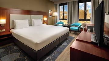 A hotel room with a large bed, two chairs, a desk with a laptop, and a TV.