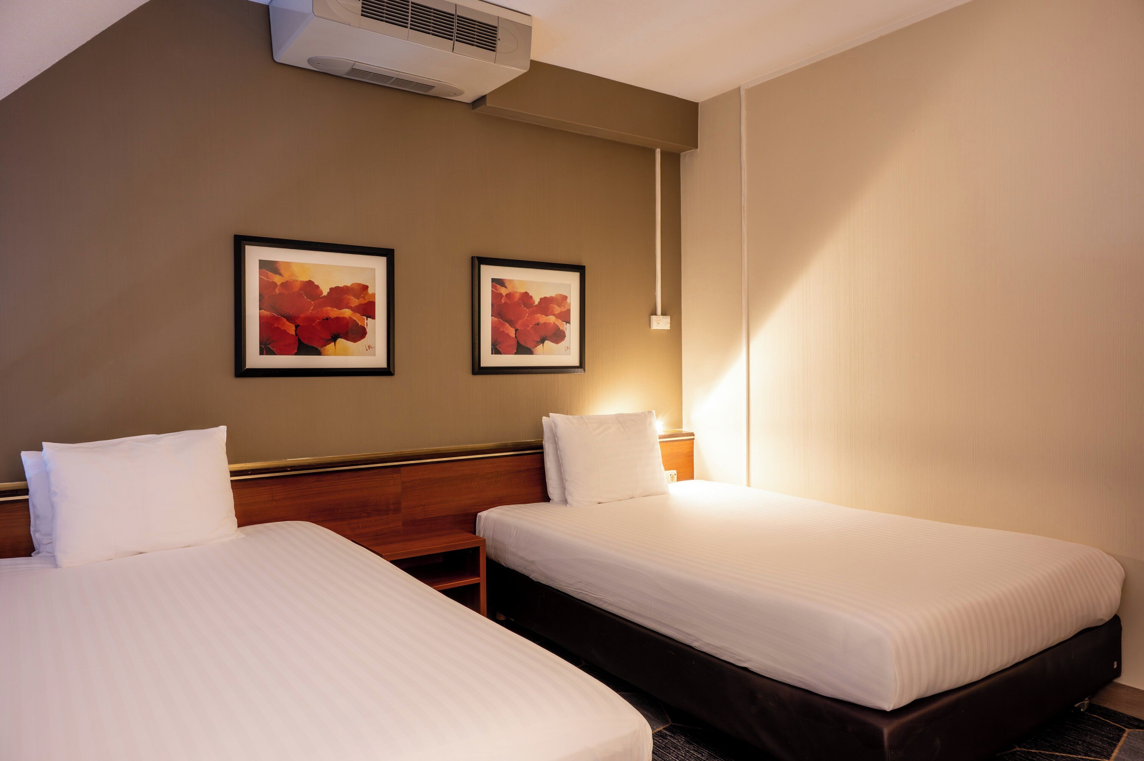A hotel room with two single beds, a wooden headboard, a wall-mounted air conditioner, and two framed pictures.