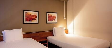 A hotel room with two single beds, a wooden headboard, a wall-mounted air conditioner, and two framed pictures.