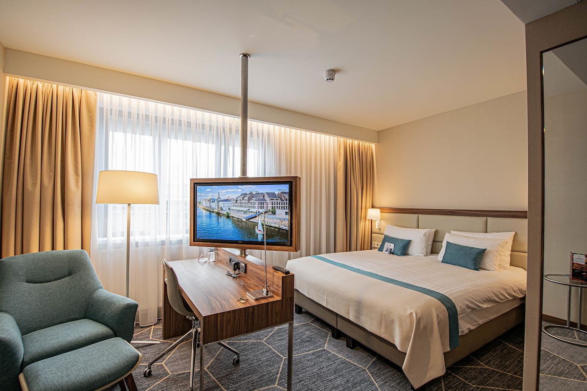 Een hotelkamer met een bed, een bureau met een computer, een stoel, een televisie en een raam met gordijnen.