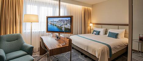 A hotel room with a bed, a desk with a computer, a chair, a television, and a window with curtains.
