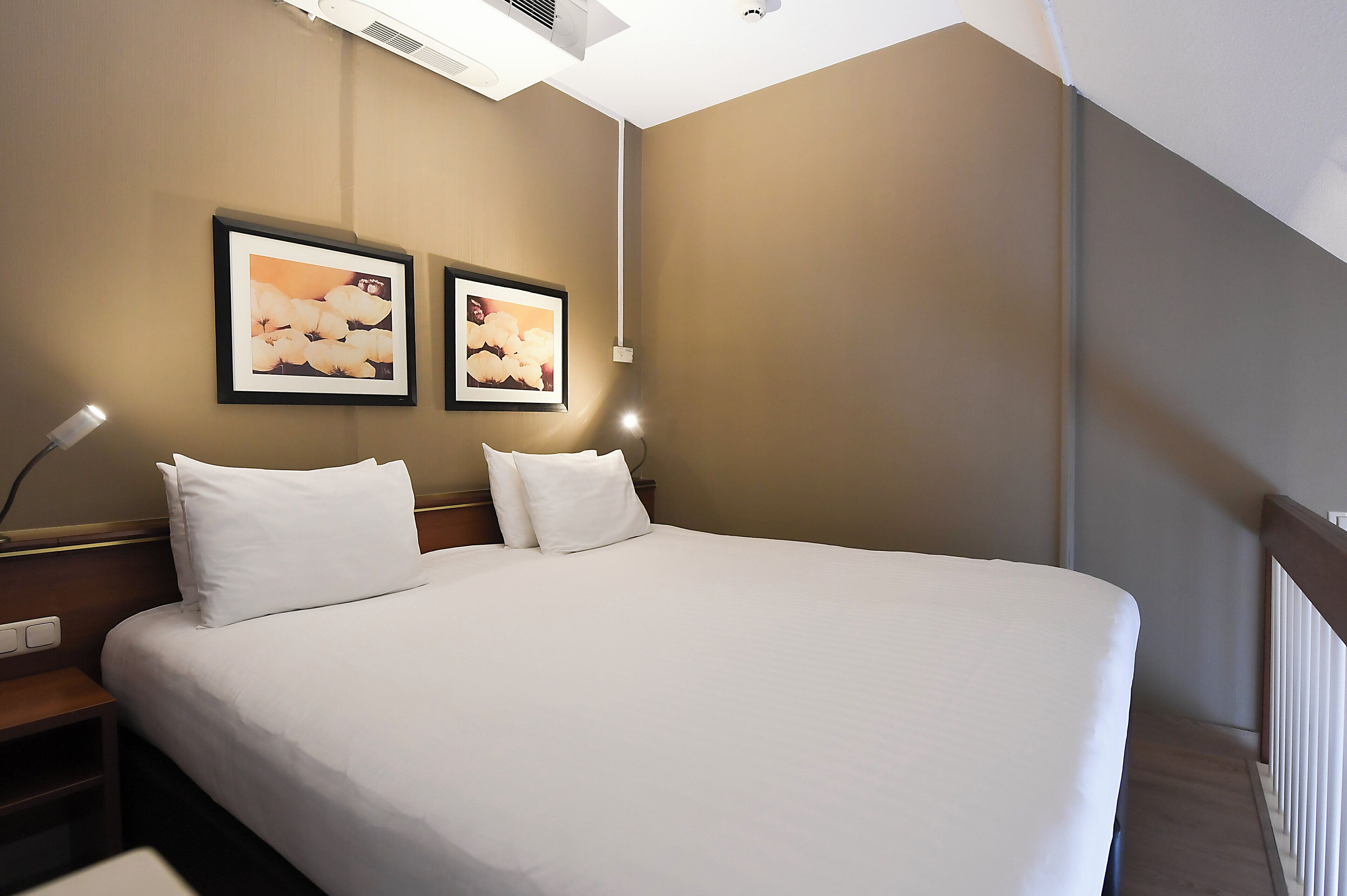 A hotel room with a large bed, two framed pictures on the wall, a nightstand, and a lamp.