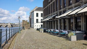 Un sentier pavé de pierres mène à un espace extérieur aménagé avec vue sur la rivière, un pont se dessine à l’arrière-plan et un bâtiment à la façade blanche se dresse.