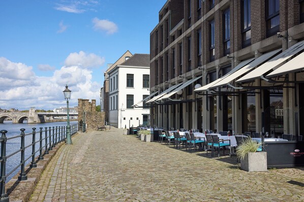 Un sentier pavé de pierres mène à un espace extérieur aménagé avec vue sur la rivière, un pont se dessine à l’arrière-plan et un bâtiment à la façade blanche se dresse.