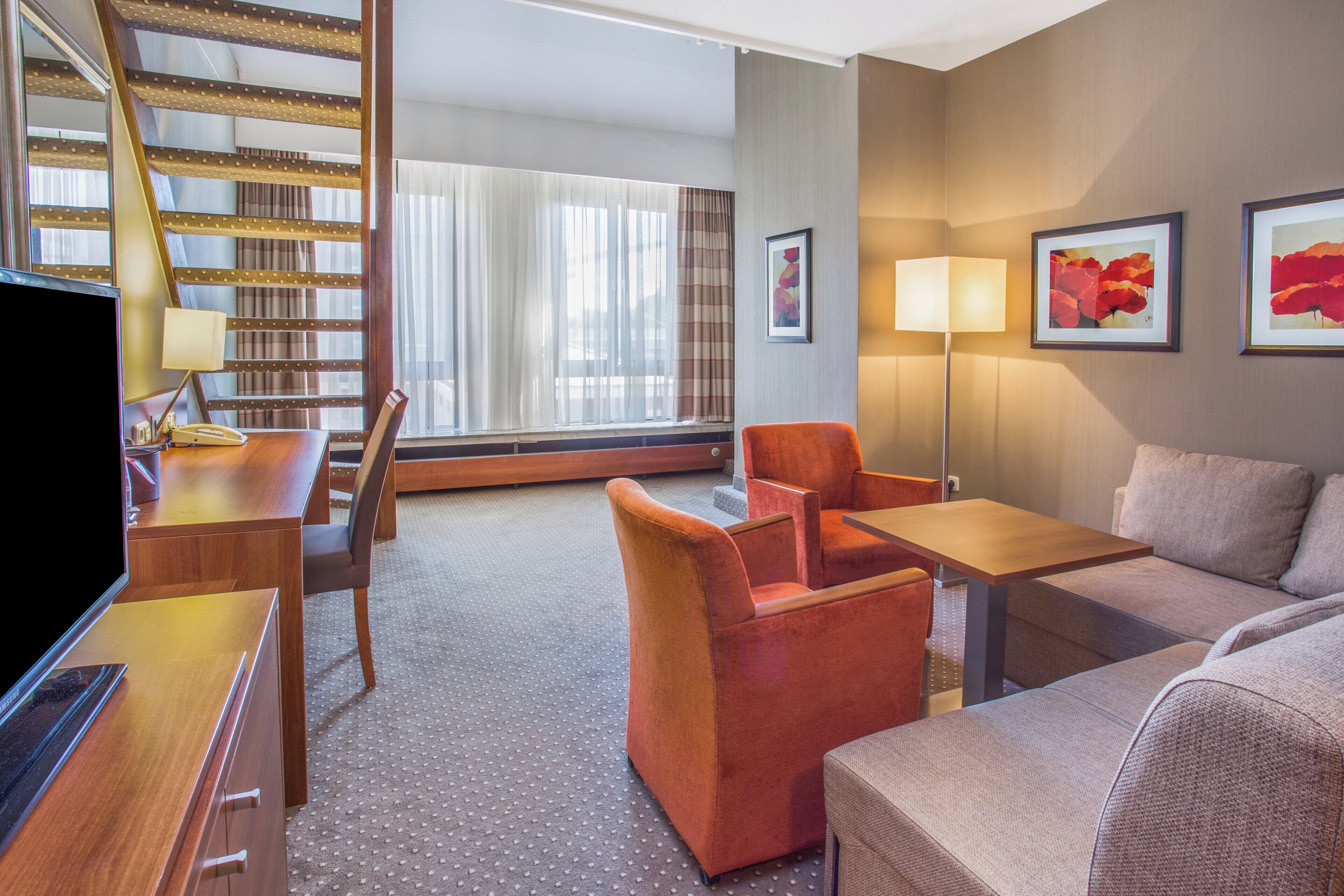 A hotel room with a sofa, armchair, a desk with a lamp, a television, and a staircase.