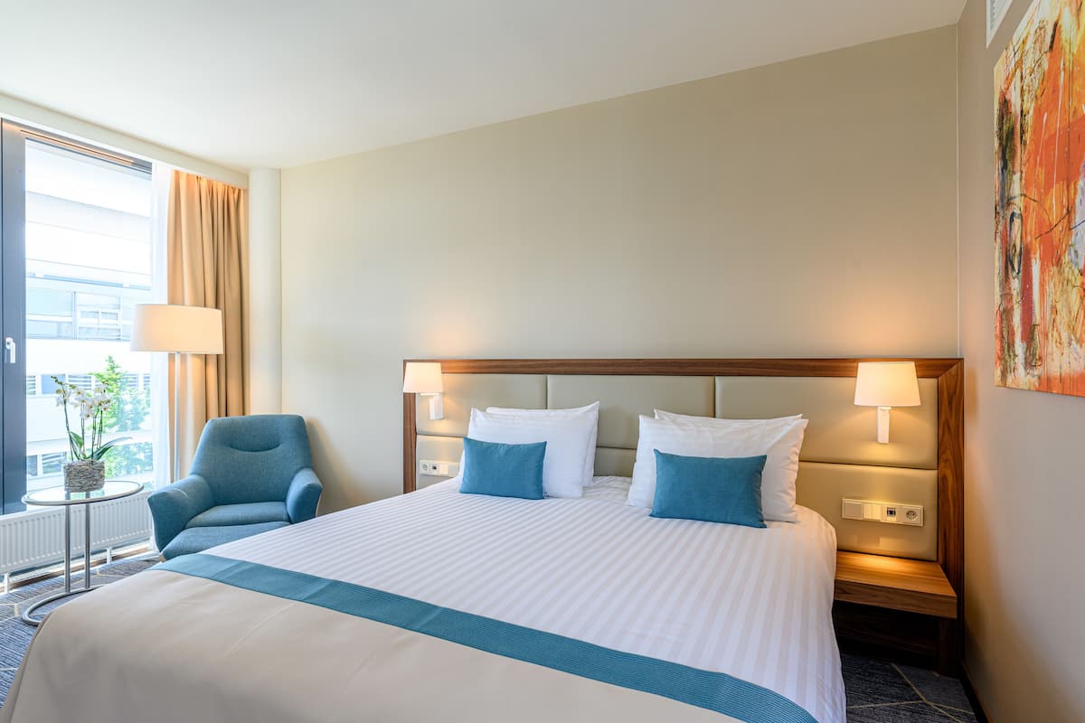 Hotelkamer met een groot bed, een stoel, een nachtkastje met een lamp en een raam met gordijnen.