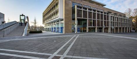 A modern building with a flat roof, large windows, and a paved plaza with a crosswalk.