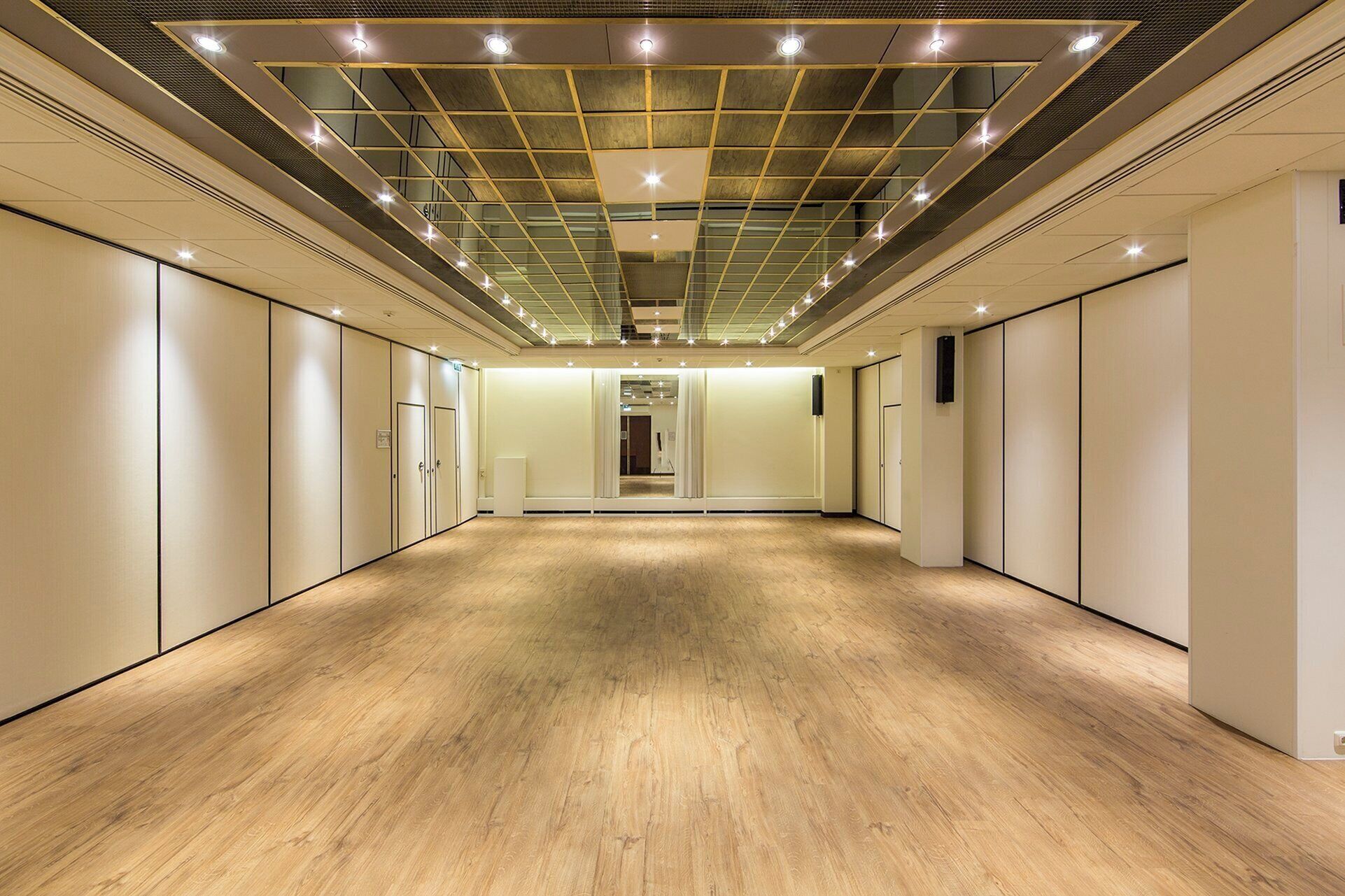 A hotel corridor with a wooden floor, mirrored walls, and a ceiling with recessed lighting.