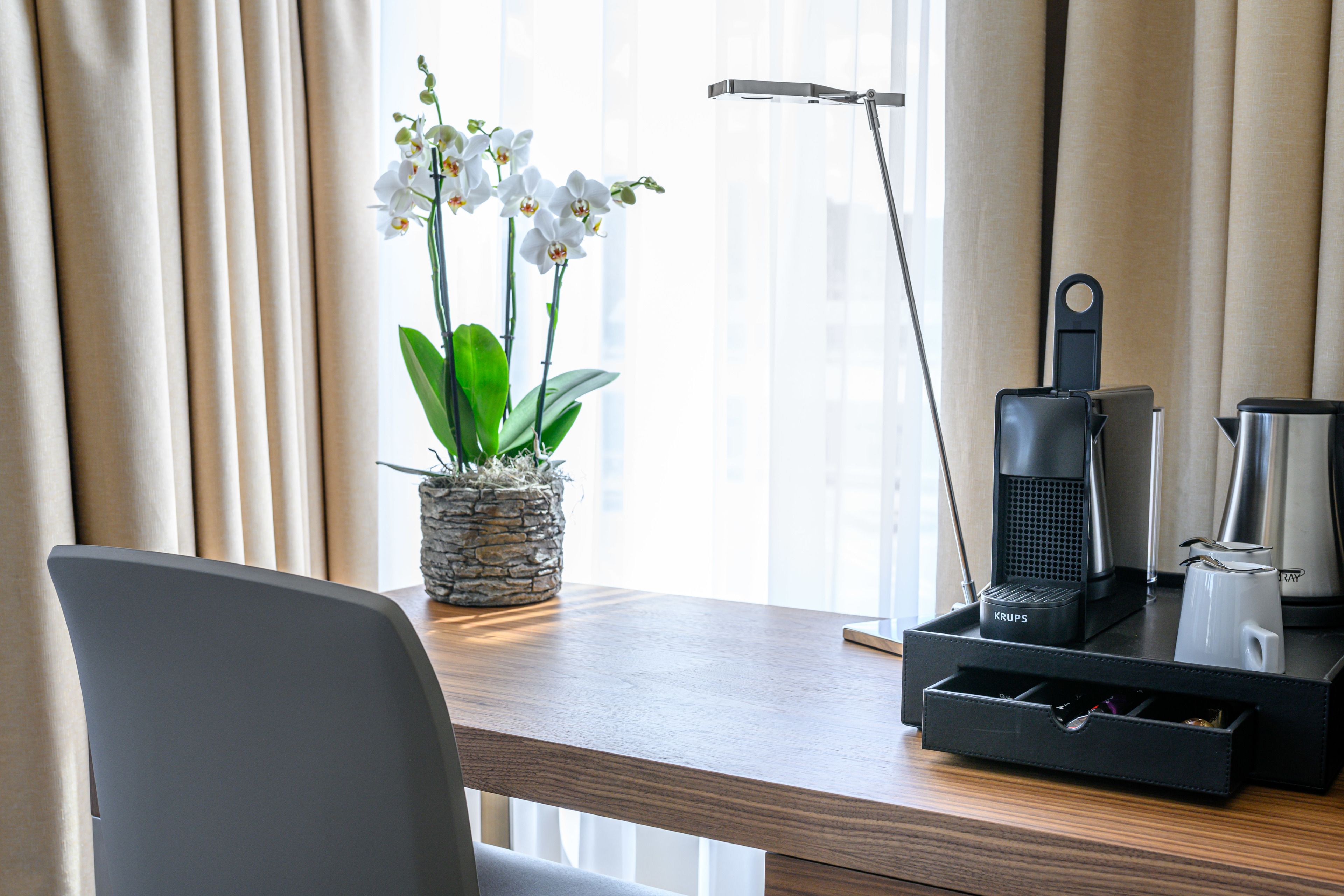 Habitación de hotel con un escritorio de madera, una orquídea en maceta, una cafetera Krups y una lámpara.