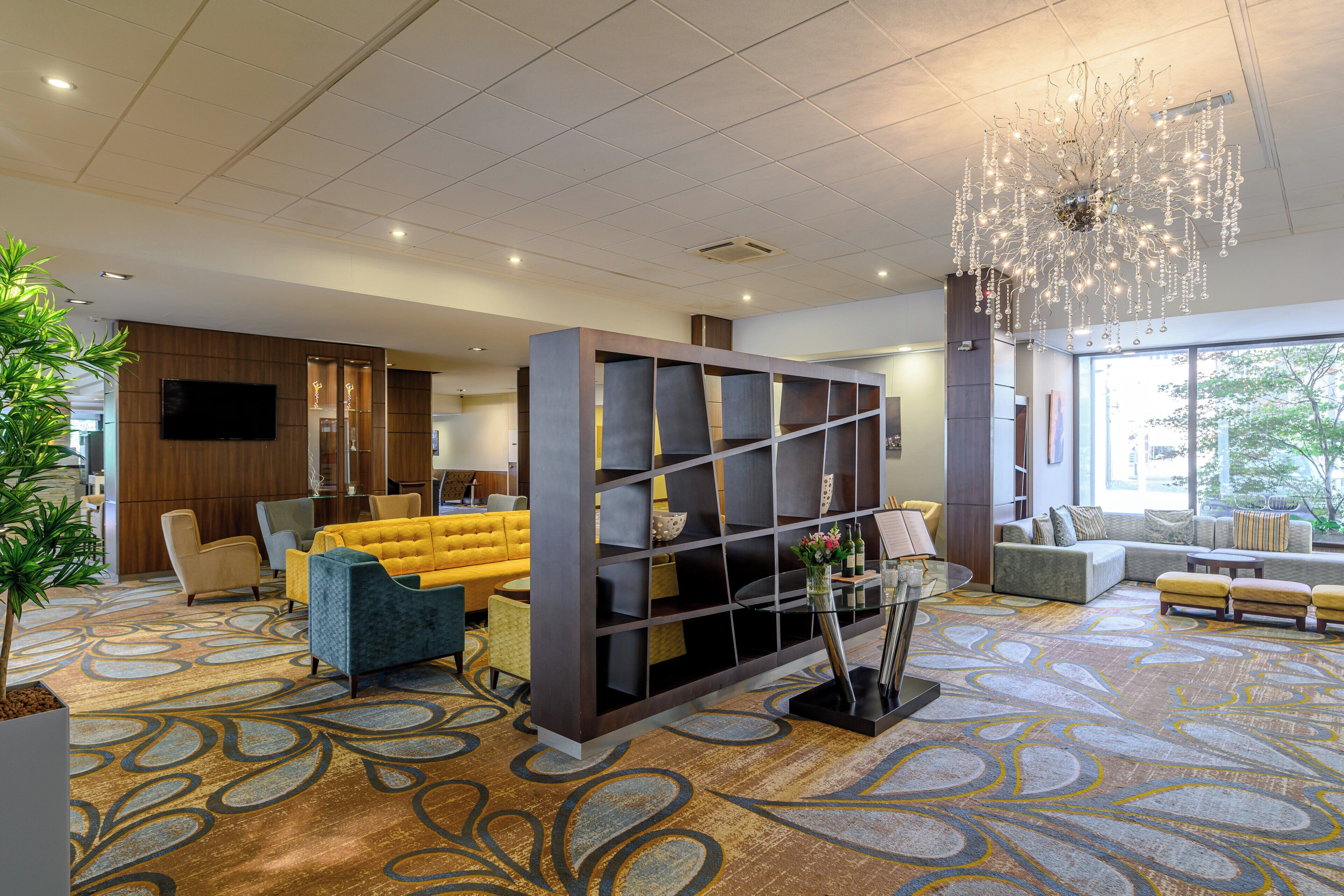 Een moderne hotellobby met een gele bank, zithoek en een decoratieve kroonluchter.