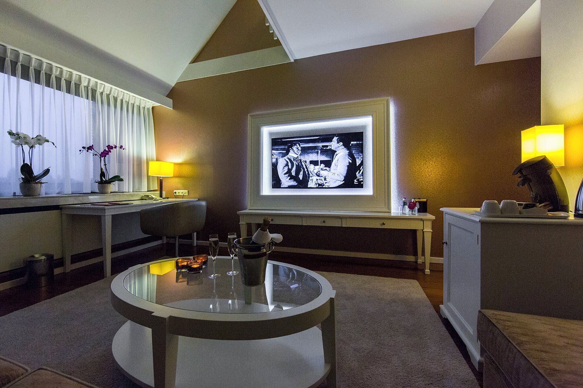 Un salon moderne avec une table basse en verre, un téléviseur à écran plat fixé au mur, un bureau avec une lampe et une orchidée en pot.