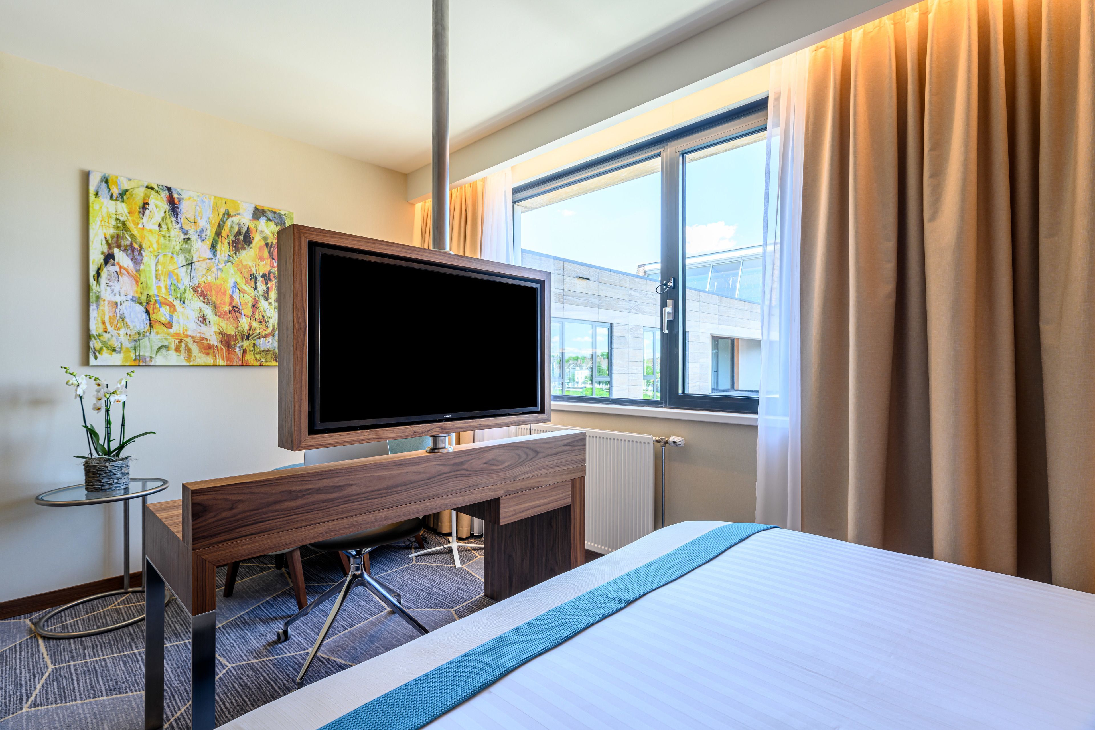 Hotelkamer met een bed, een tv op een houten standaard, een kleintje tafeltje met een plant en een raam met gordijnen.