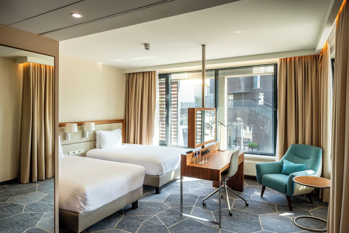 Ein Hotelzimmer mit zwei Betten, einem Schreibtisch mit Stuhl, einem kleinen Tisch und einem großen Fenster mit Vorhängen.