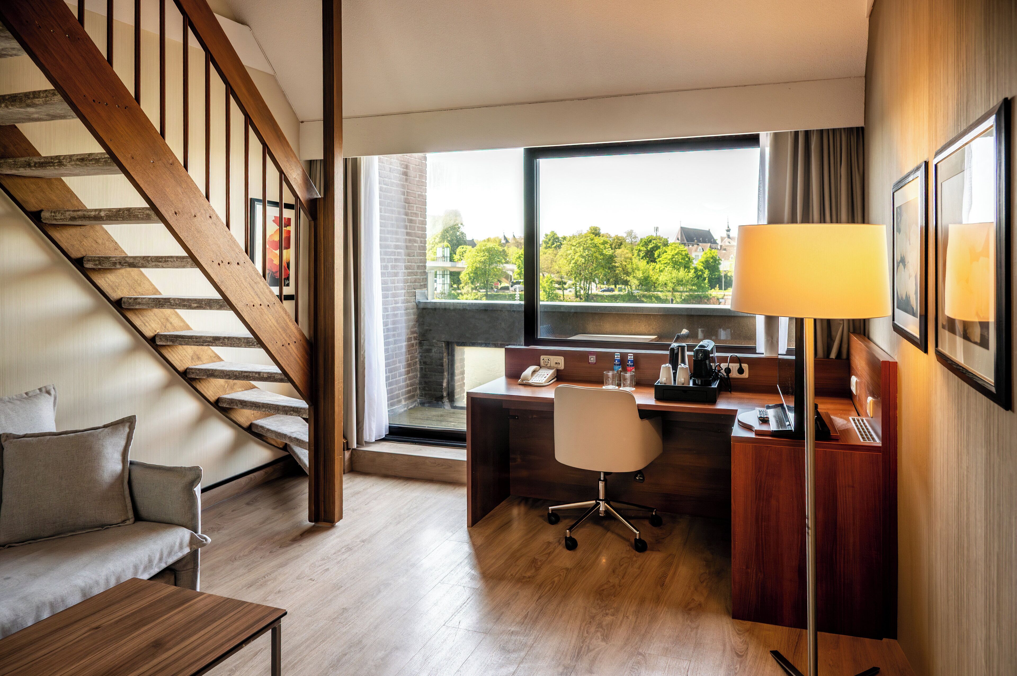 A hotel room with a wooden staircase, a desk with a chair, a sofa, and a large window offering a city view.