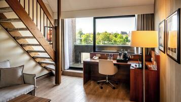 A hotel room with a wooden staircase, a desk with a chair, a sofa, and a large window offering a city view.