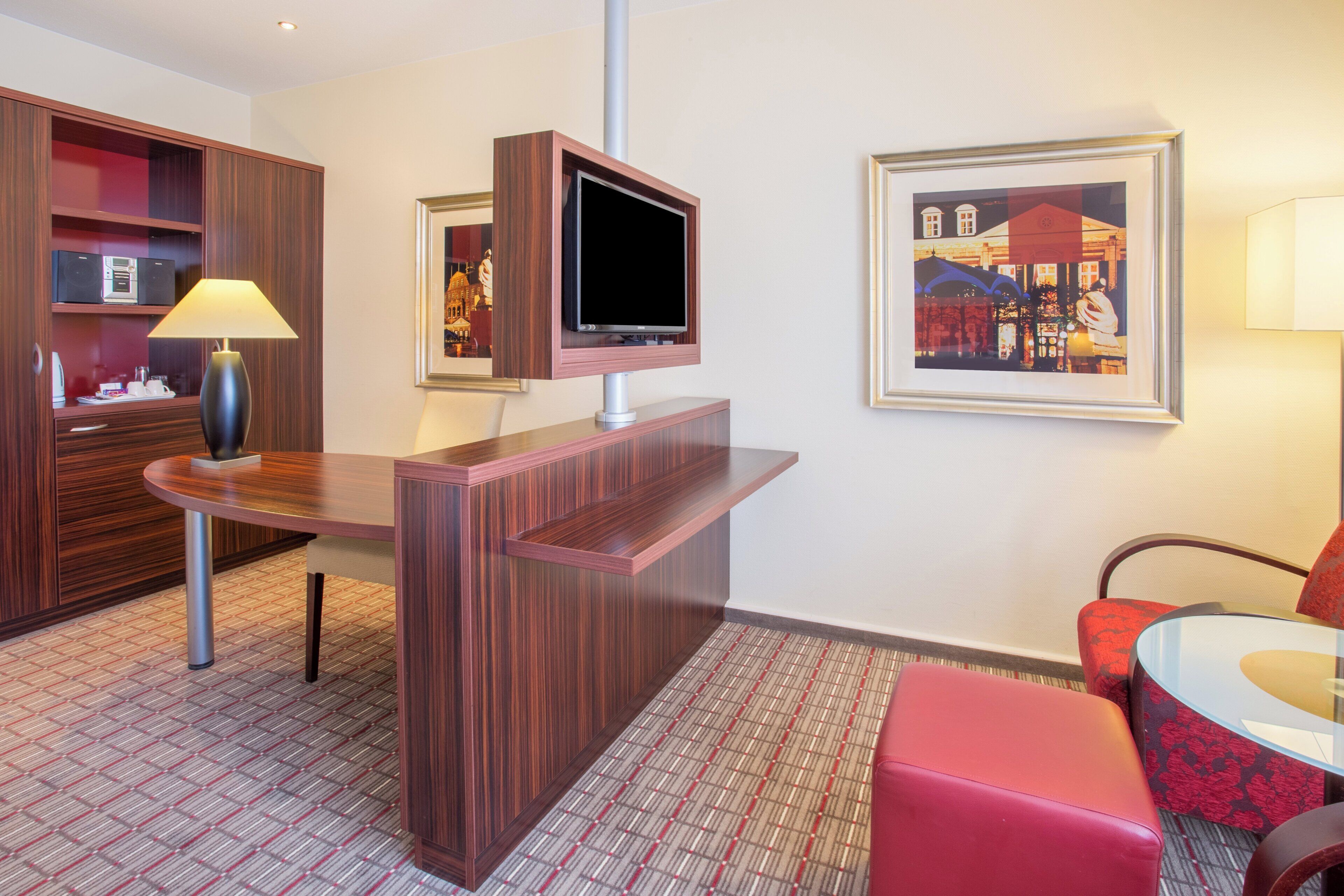 A hotel room with a wooden desk, a flat-screen TV mounted on the wall, a red armchair, and a framed picture on the wall.