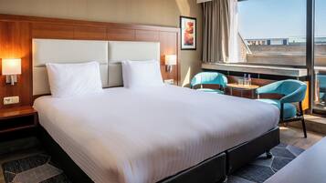 Quarto de hotel com uma cama grande, duas luminárias de cabeceira, uma mesinha com cadeiras e uma janela com vista para prédios.