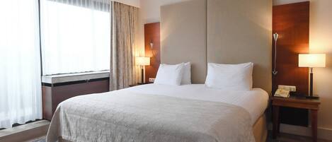 Habitación de hotel con una cama grande, dos mesitas de noche con lámparas, una ventana con cortinas y un cabecero de madera.