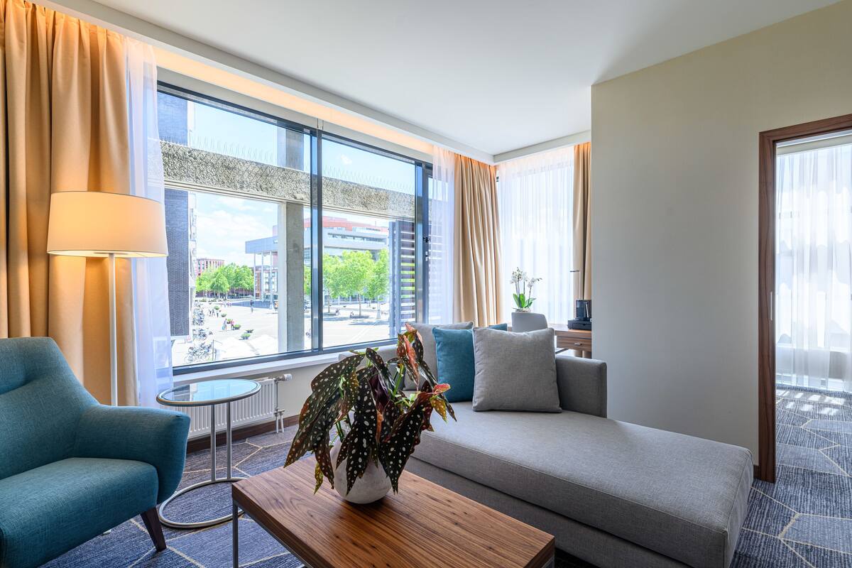 A hotel room with a sofa, armchair, and a small table with a plant. There is a large window with curtains, a view of the street outside, and a potted plant on the windowsill.