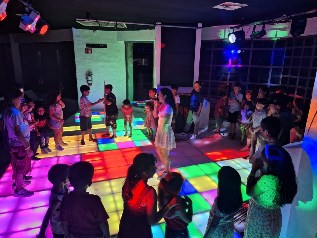 Pista de dança com iluminação colorida, rodeada de pessoas.