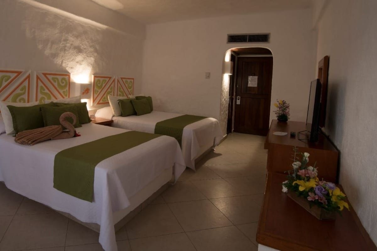A hotel room with two beds, a wooden desk, a television, and a small table with flowers.