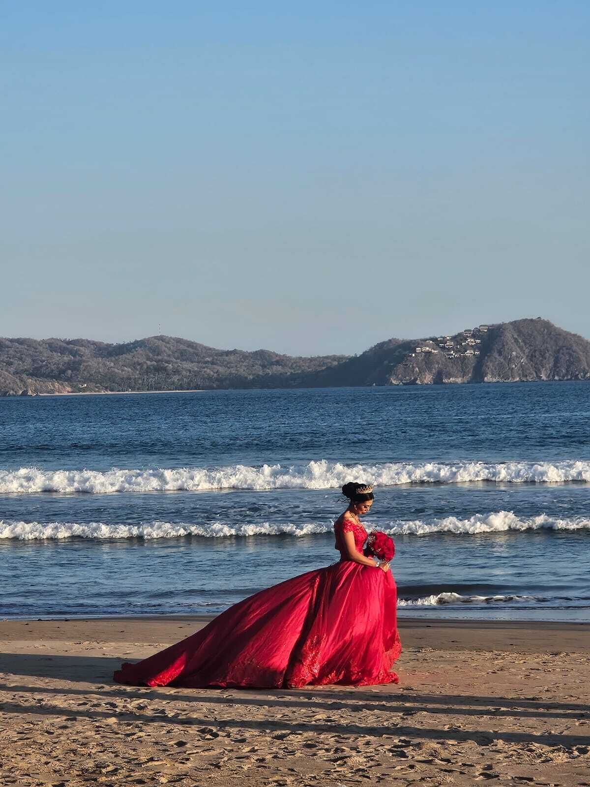 Una playa con olas, una persona con un vestido rojo y una colina distante con edificios.