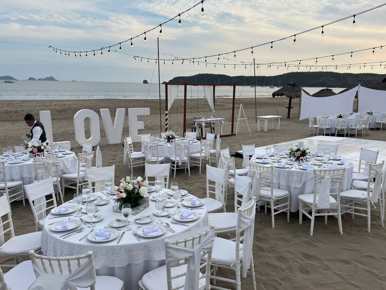 Montaje para una boda al aire libre con mesas redondas, sillas blancas y luces colgantes en una playa.